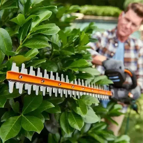 WG259E-WORX-CORDLESS-HEDGE-TRIMMER-20V-61CM-2 Close-up of orange hedge trimmer blade cutting green plant leaves with gardener working in background